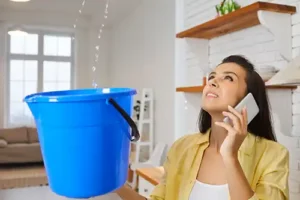 At Your Service Roofing—An unhappy female homeowner holds a blue bucket to catch the water from her leaky roof as she talks on the phone to a roofer.