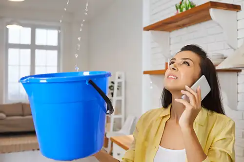 At Your Service Roofing—An unhappy female homeowner holds a blue bucket to catch the water from her leaky roof as she talks on the phone to a roofer.